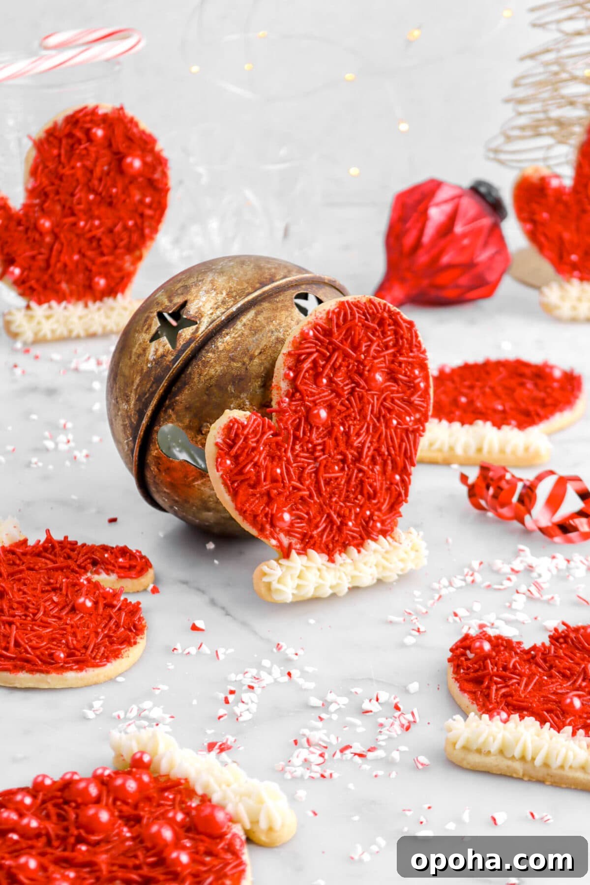 Santa’s Glove Peppermint Sugar Cookies: Festive Holiday Recipe 5 seven glove shaped cookies, with one leaning against a red bell in the middle on marble surface with crushed peppermint around.