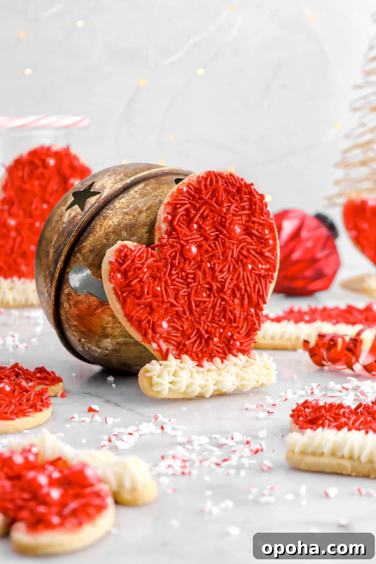 Santa’s Glove Peppermint Sugar Cookies: Festive Holiday Recipe 6 glove cookie leaning against bell with more cookies around, a red ornament, and fairy lights behind.