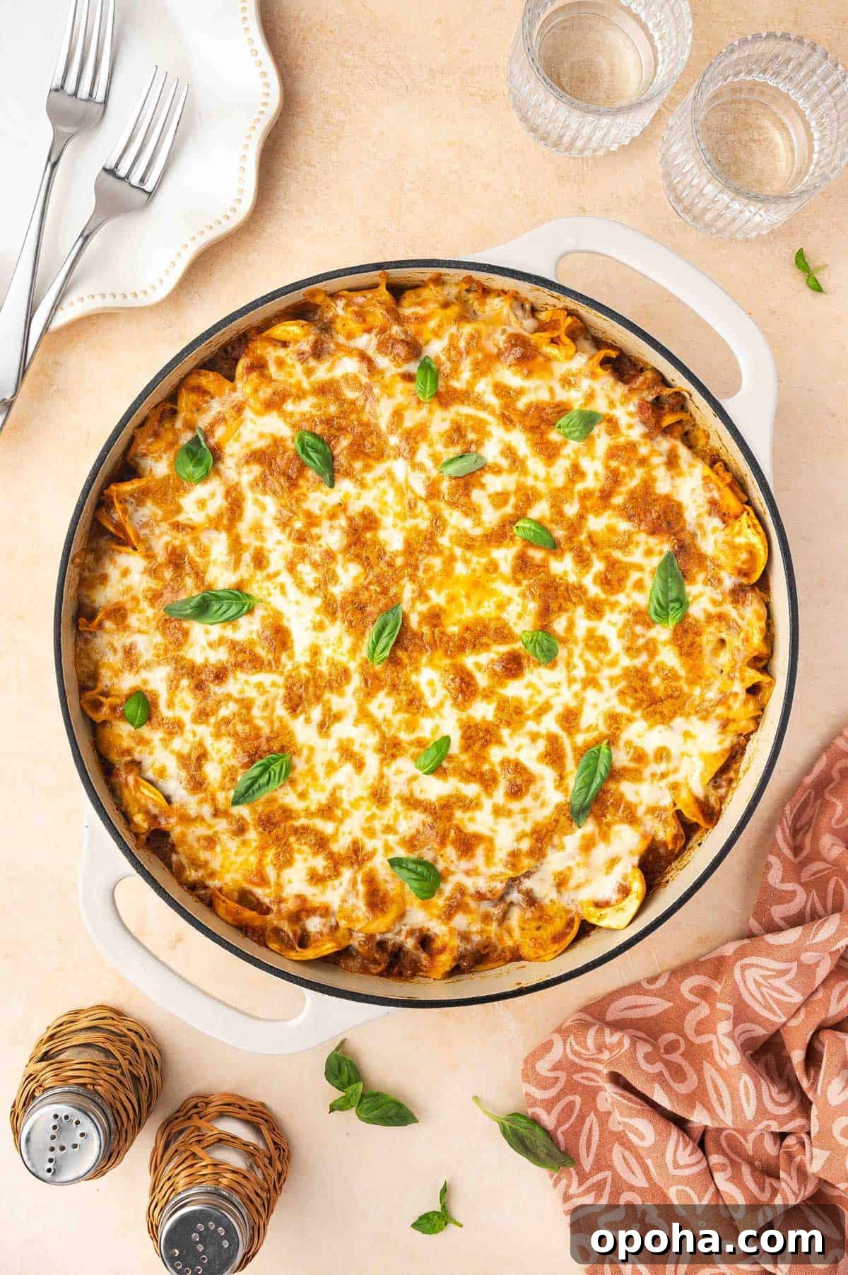 Tortellini Lasagna Bake: Cheesy Italian Pasta Casserole Recipe 9 Overhead view of a skillet of tortellini lasagna with basil garnish. Linen napkin on the side with drinks, salt and pepper shakers, and forks on a plate.