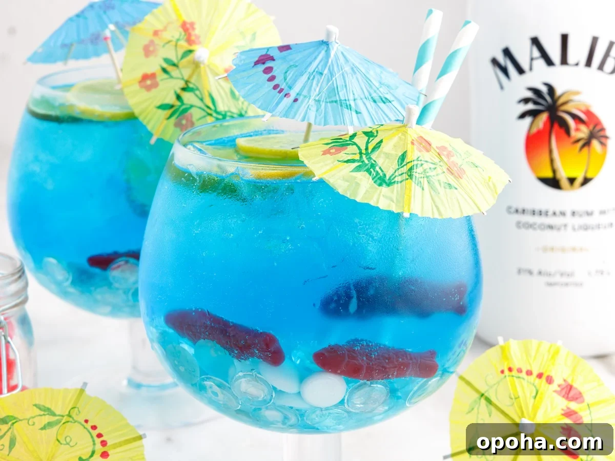 Two large round glasses filled with bright blue Fish Bowl Drink cocktails, ice, lime slices, and gummy fish, each topped with colorful paper umbrellas and striped straws. A bottle of Malibu rum is in the background.