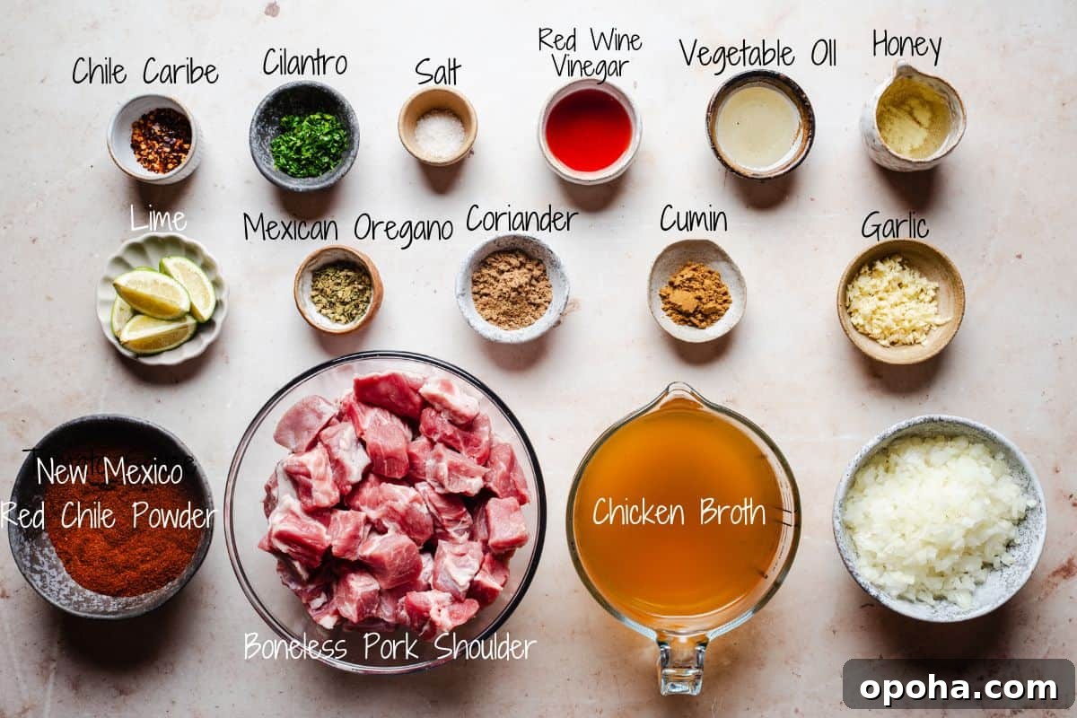 Authentic New Mexican Carne Adovada Recipe 3 Ingredients for a Carne Adovada recipe are displayed on a table, including chile caribe, cilantro, lime, spices, and boneless pork shoulder. Other ingredients like red wine vinegar, honey, chicken broth, garlic, and diced onion are also shown.