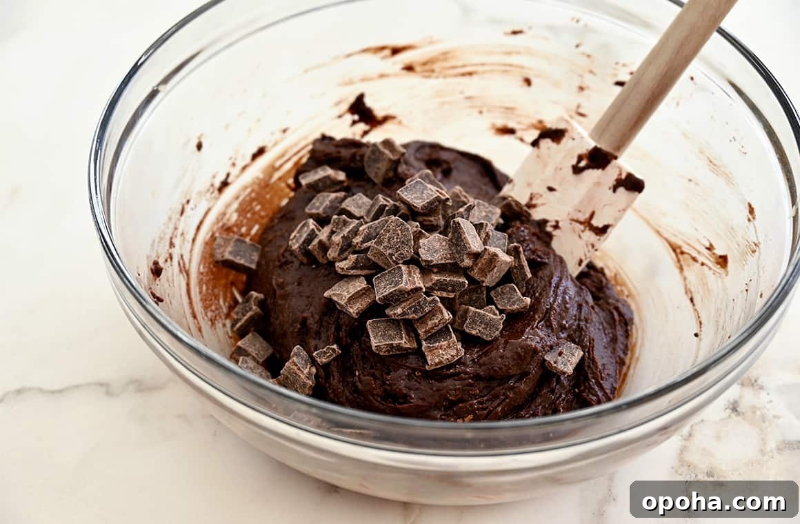 Fudgy From-Scratch Brownie Cupcakes Recipe 4 A clear bowl containing brownie cupcake batter with chocolate chunks and a spatula.