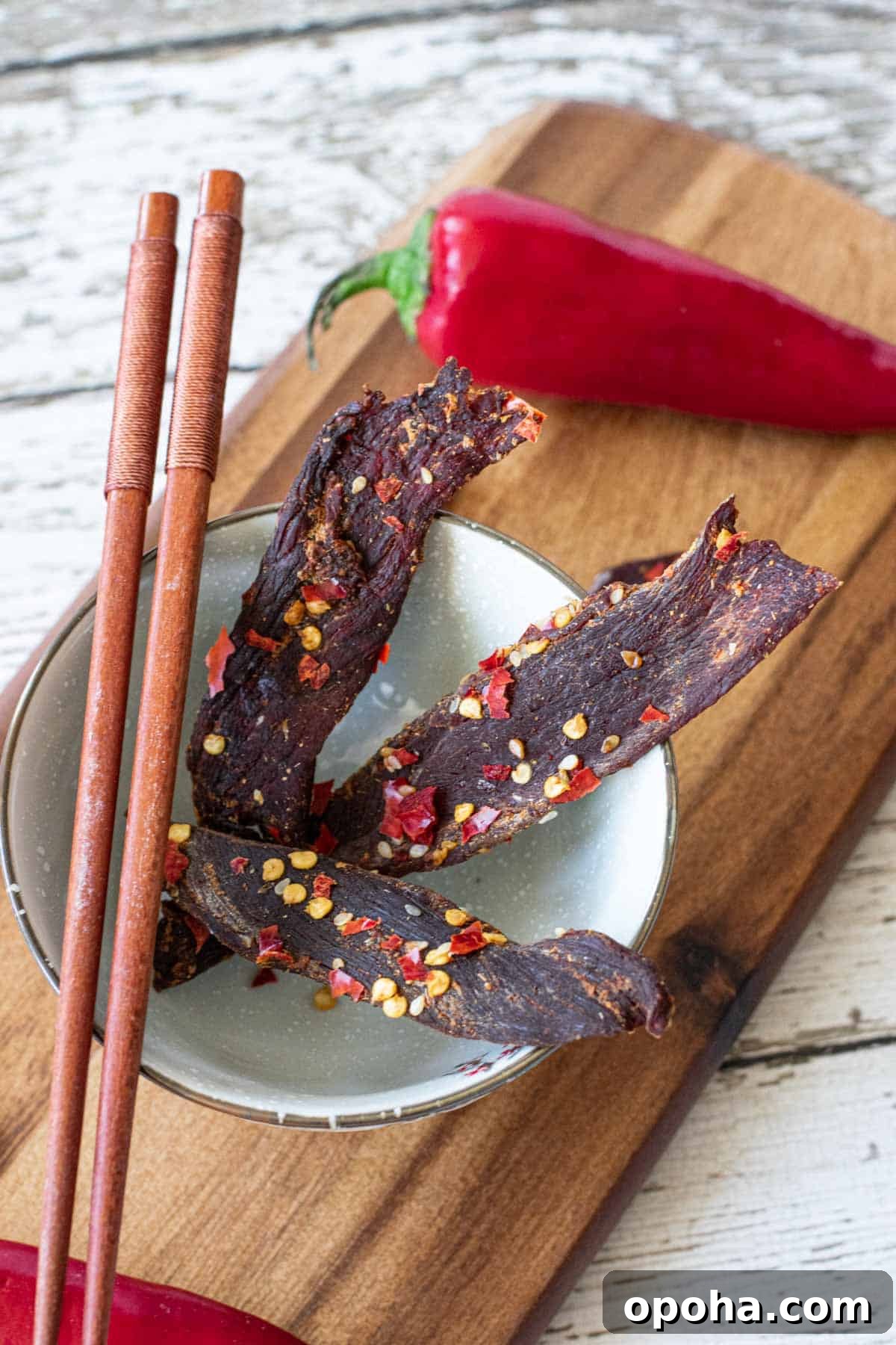 Sesame-Glazed Venison Jerky Recipe: Savory Sweet Game Meat Snack 2 venison jerky in bowl atop a cutting board with chop sticks and Fresno chili