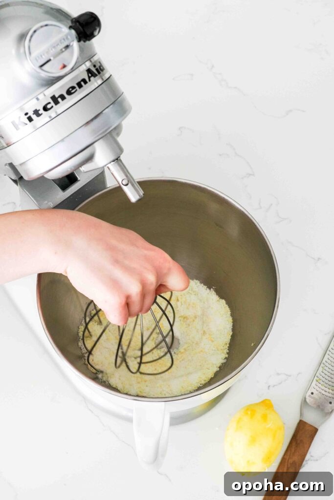 Zesty Lemon Madeleines: Classic French Tea Cakes Recipe 5 A hand uses the whisk on a KitchenAid mixer to stir together lemon zest and sugar.