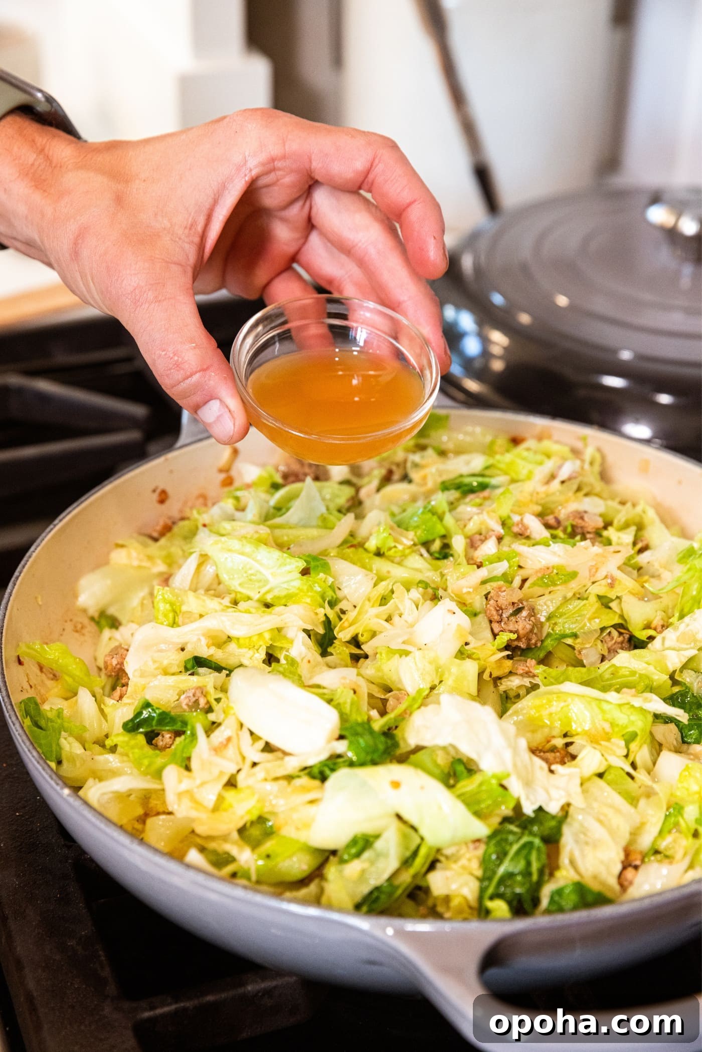 One-Pan Skillet Cabbage with Sausage and Garlic Butter 10 Hand holding small glass bowl filled with brown-colored apple cider vinegar about to pour it on top of sliced cabbage in large skillet.