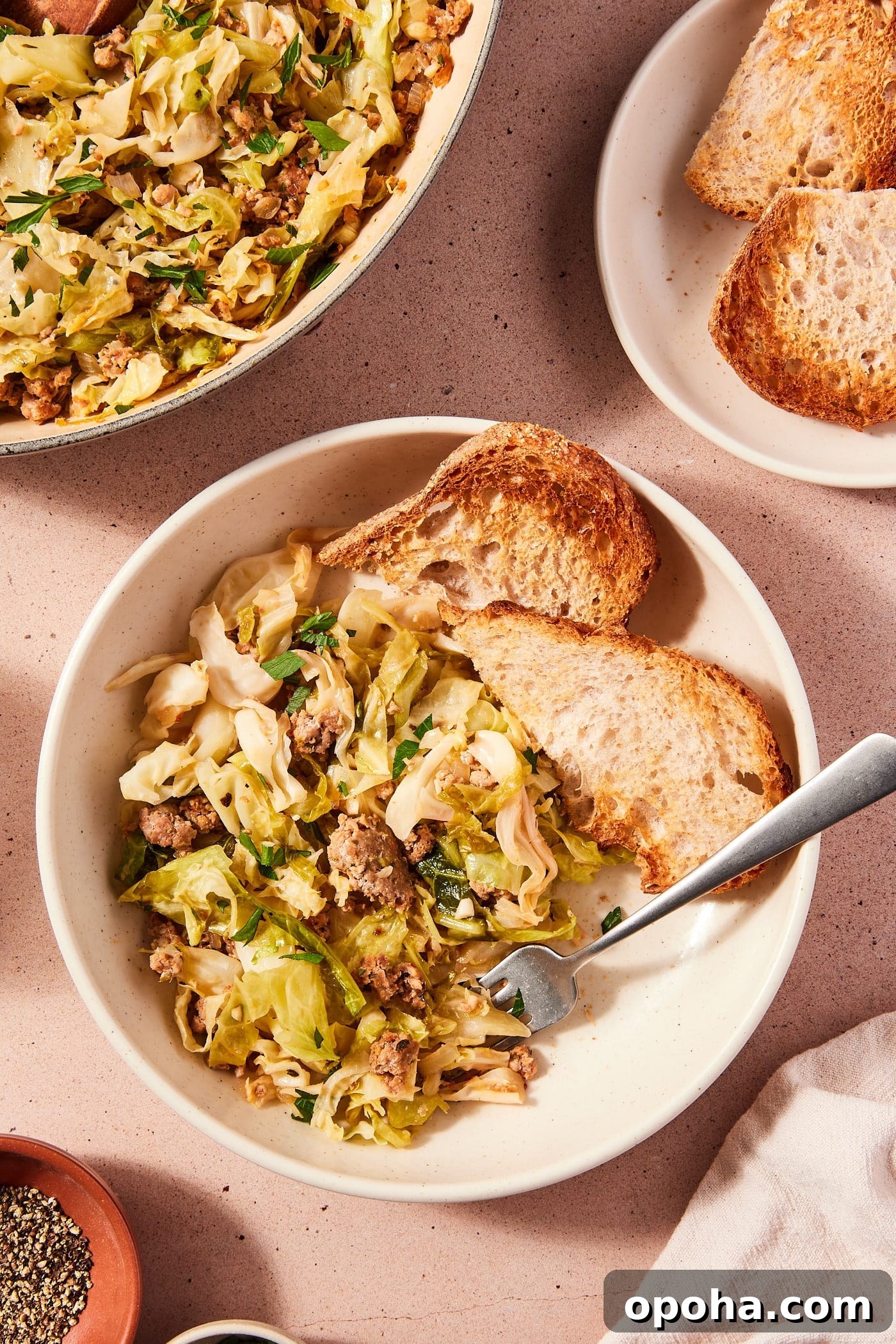 One-Pan Skillet Cabbage with Sausage and Garlic Butter 12 White bowl filled with serving of skillet sausage and cabbage with slices of toasted bread alongside with skillet filled with rest of the cooked cabbage.