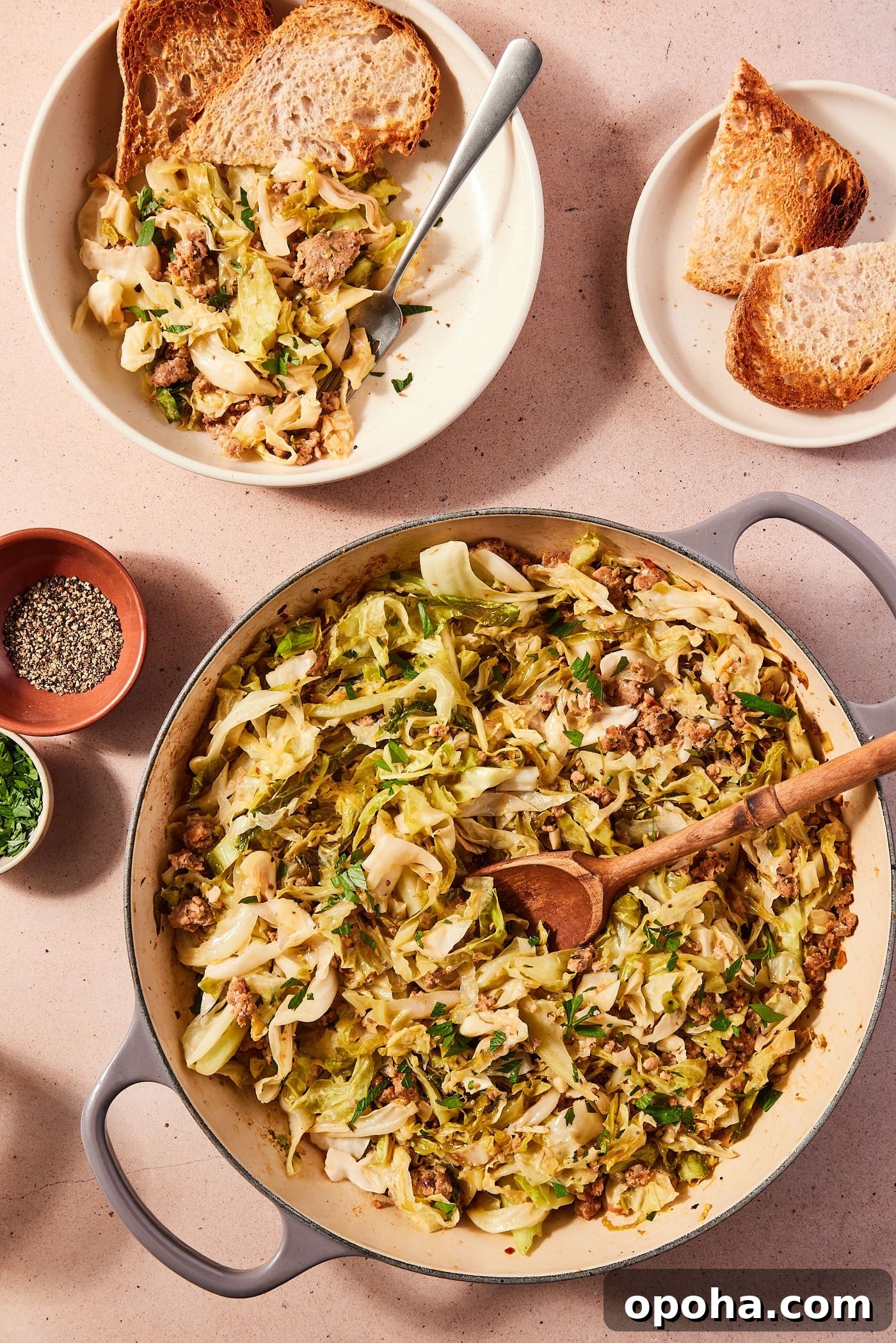 One-Pan Skillet Cabbage with Sausage and Garlic Butter 3 Large skillet filled with cooked cabbage and sausage with wooden spoon scooping up a serving with a serving already in a bowl with toasted slices of bread.