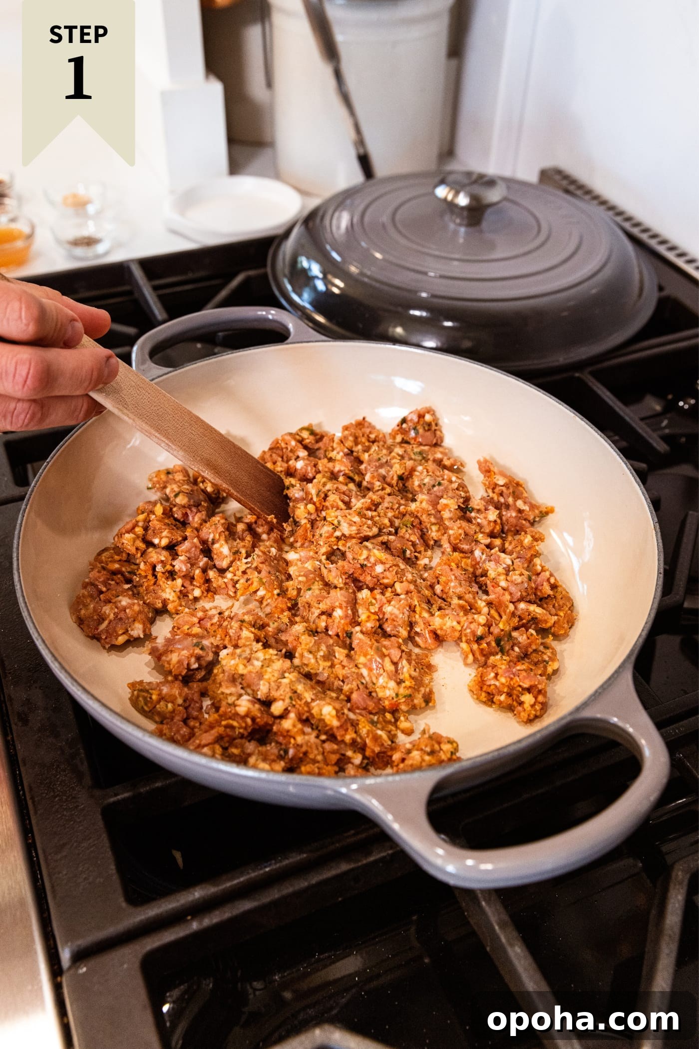 One-Pan Skillet Cabbage with Sausage and Garlic Butter 5 Hand holding wooden spoon breaking up ground sausage in large skillet on stovetop with slight browning.