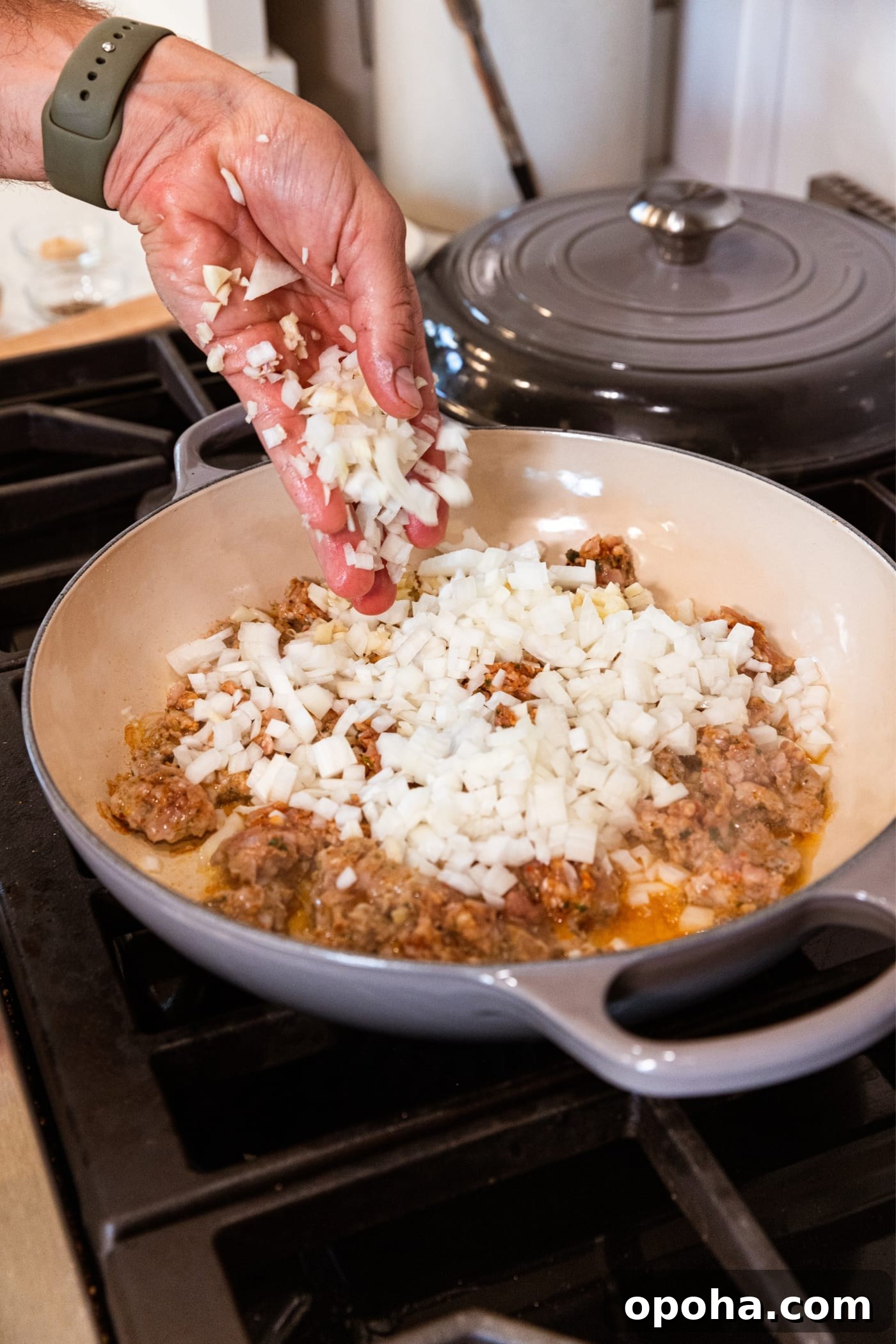 One-Pan Skillet Cabbage with Sausage and Garlic Butter 6 Hand pouring diced onion into large gray skillet on stovetop with cooking ground sausage underneath.