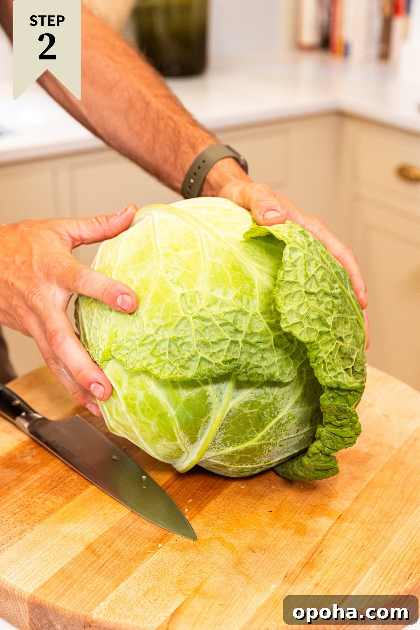 One-Pan Skillet Cabbage with Sausage and Garlic Butter 7 Hand holding large whole light green colored cabbage on wooden cutting board before slicing with knife.