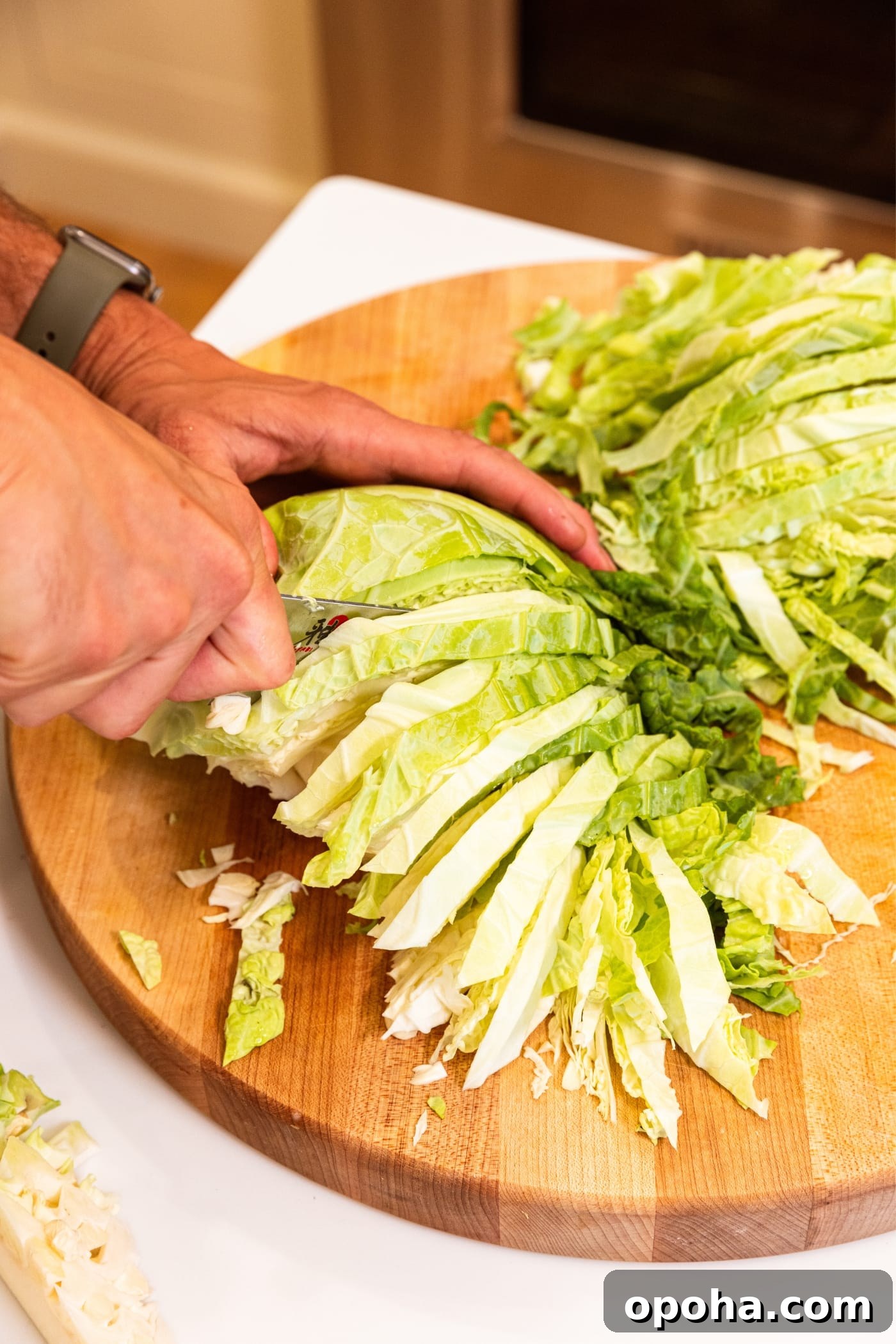 One-Pan Skillet Cabbage with Sausage and Garlic Butter 8 Hand using large knife to slice light green colored cabbage into small slices on wooden cutting board.