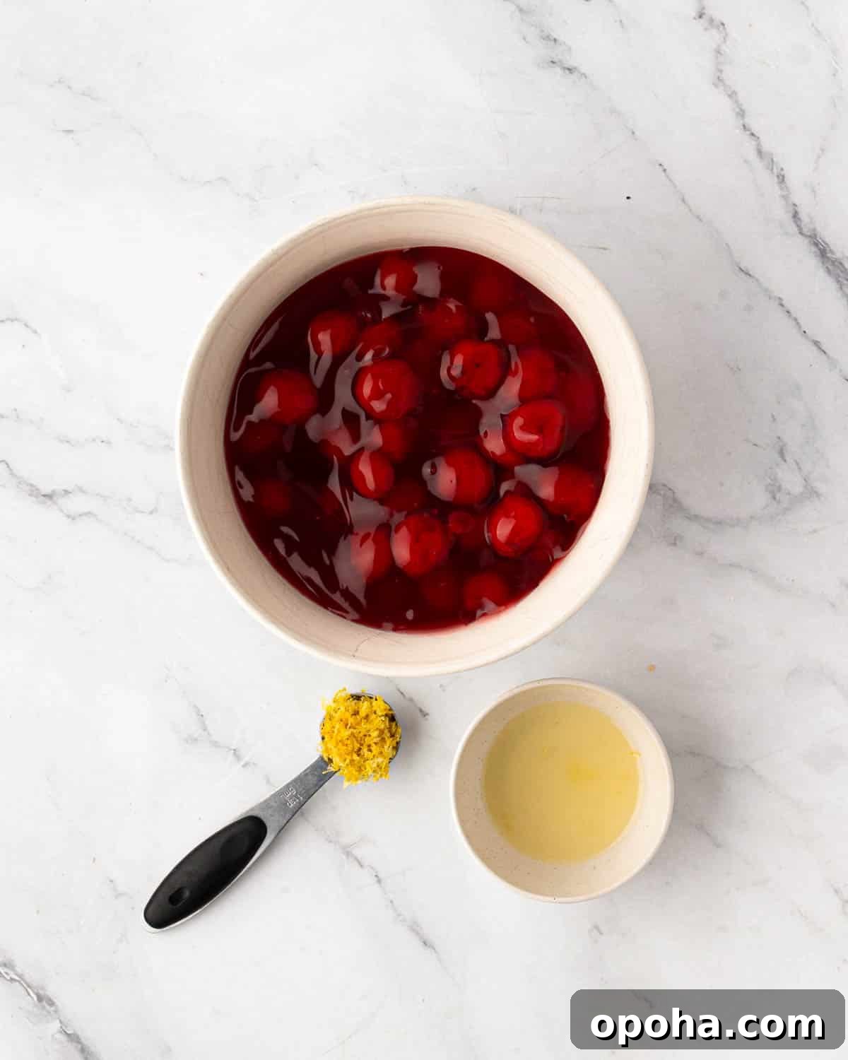 Cherry Pie Bars Recipe: Easy Homemade Cherry Dessert Bars 13 A white bowl with cherry pie filling, a measuring spoon with lemon zest, and lemon juice in a small white bowl.