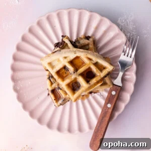 Overhead image of chocolate chip waffles cut into quarters and stacked on a pink scalloped plate with syrup.