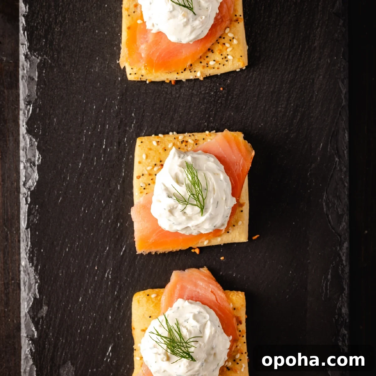Three square crackers topped with smoked salmon and cream cheese on a black stone platter.