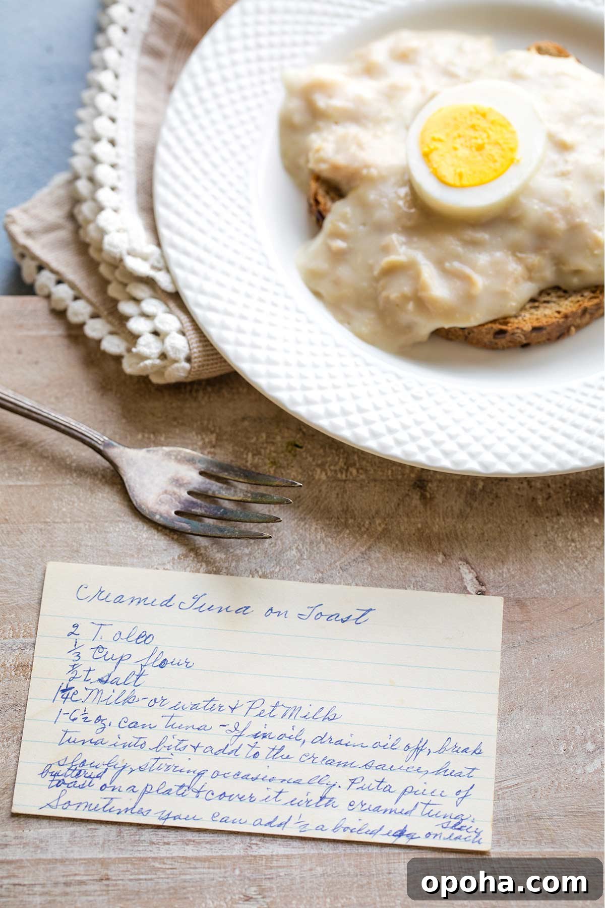 My Grandma's Creamed Tuna on Toast recipe card on a cutting board in front of her variation of serving it with a hard-boiled egg.