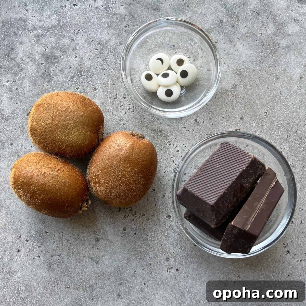 Three whole kiwis, chocolate pieces in a glass bowl, and candy eyes in another bowl are arranged on a gray surface.