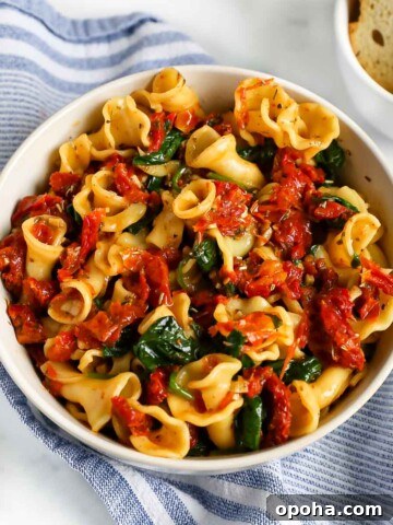 There is a white bowl filled with sun dried tomatoes, baby spinach, spices and herbs over cooked pasta.