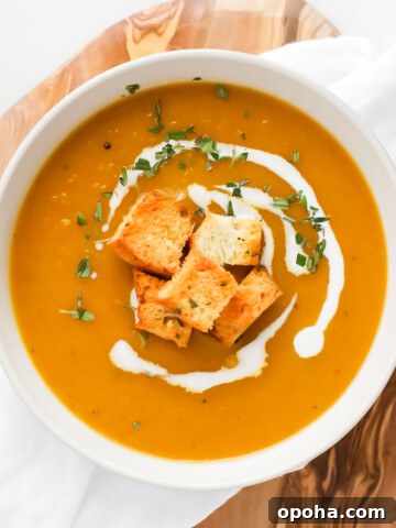A white bowl filled with orange soup garnished with coconut milk, croutons, and herbs.