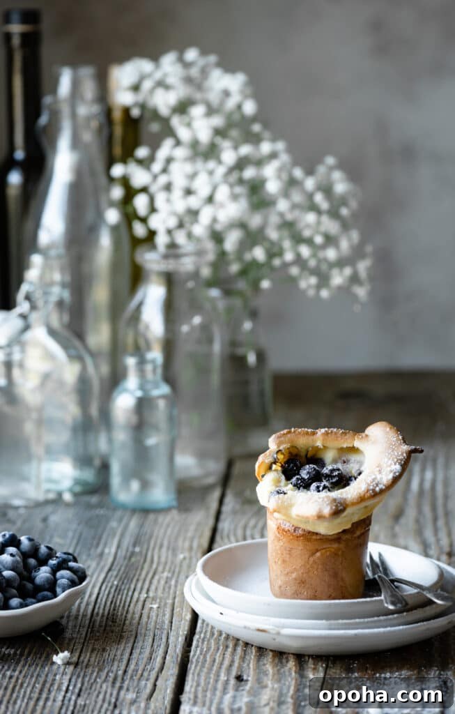Fluffy Blueberry Popovers: Light, Crispy Breakfast Treat 2 Single popover on a stack of plates on wood table next to flowers and a bowl of blueberries.