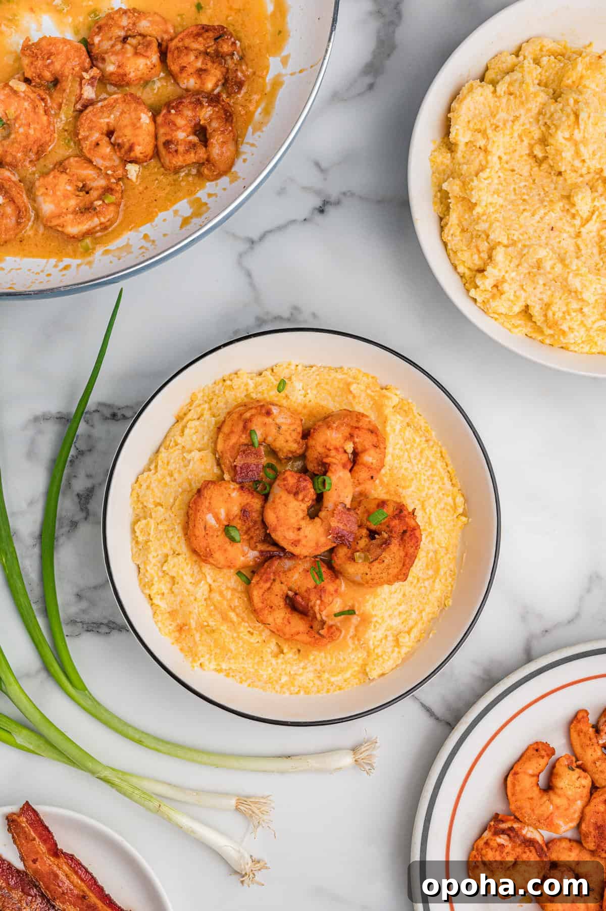 Creamy Shrimp and Grits with Cheddar and Garlic 2 Overhead view of a bowl of cheesy shrimp and grits with bacon and green onions. Surrounding it are a skillet with saucy shrimp, a bowl of cheesy grits, a plate of cooked shrimp, fresh green onions, and a plate of bacon, all arranged on a marble surface.