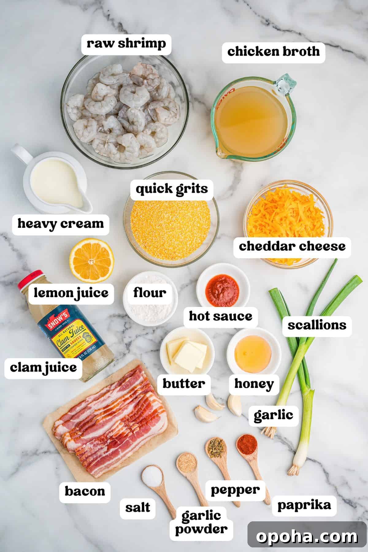 Creamy Shrimp and Grits with Cheddar and Garlic 4 Overhead view of various labeled ingredients for cheesy shrimp and grits arranged on a marble surface. Items include raw shrimp, chicken broth, quick grits, cheddar cheese, heavy cream, lemon juice, flour, hot sauce, butter, honey, garlic, scallions, bacon, clam juice, and seasonings in wooden spoons: salt, garlic powder, pepper, and paprika.