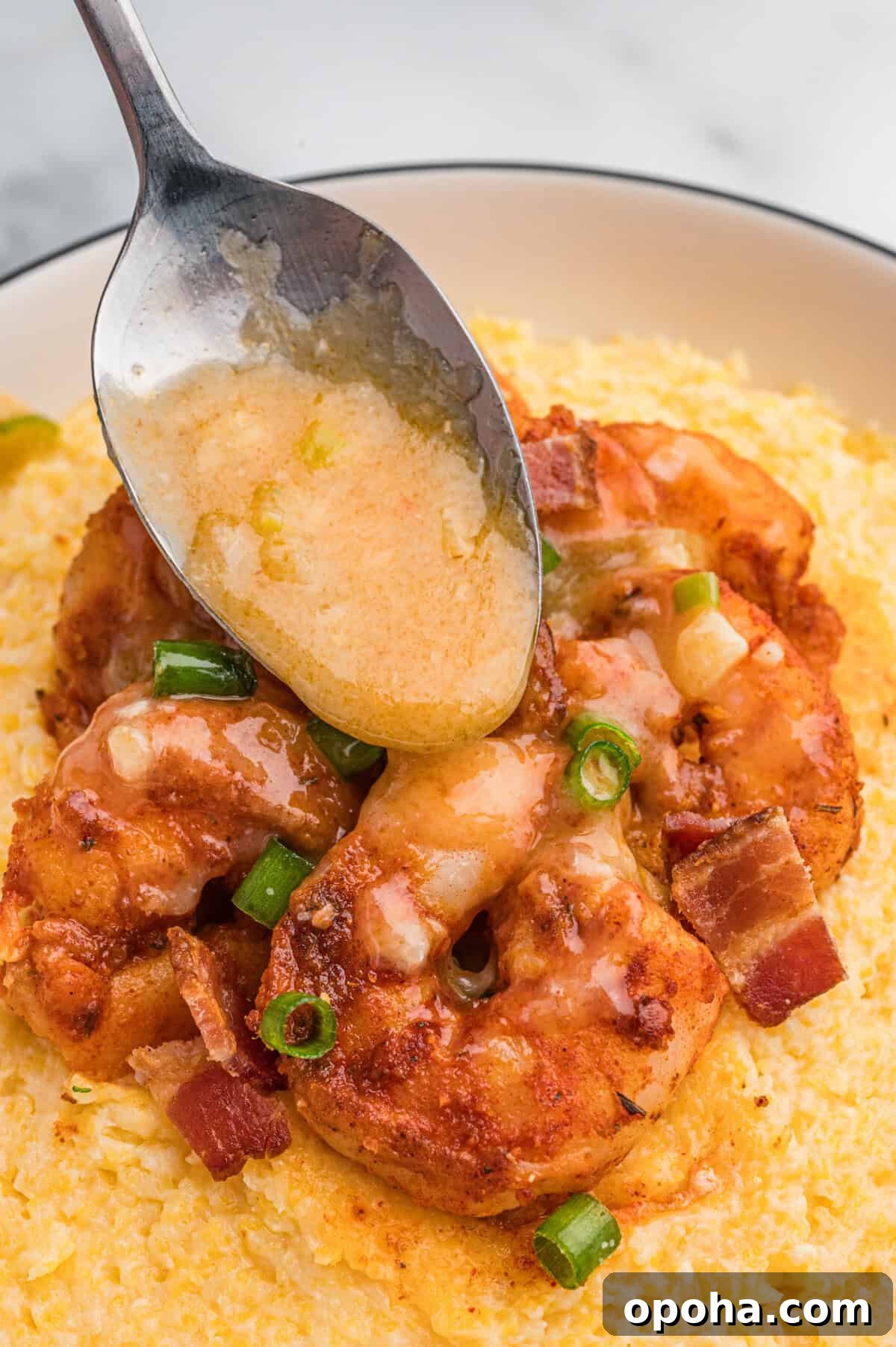 Creamy Shrimp and Grits with Cheddar and Garlic 10 A spoon drizzles buttery sauce over a serving of shrimp on a bed of cheesy grits, garnished with bacon and green onions.