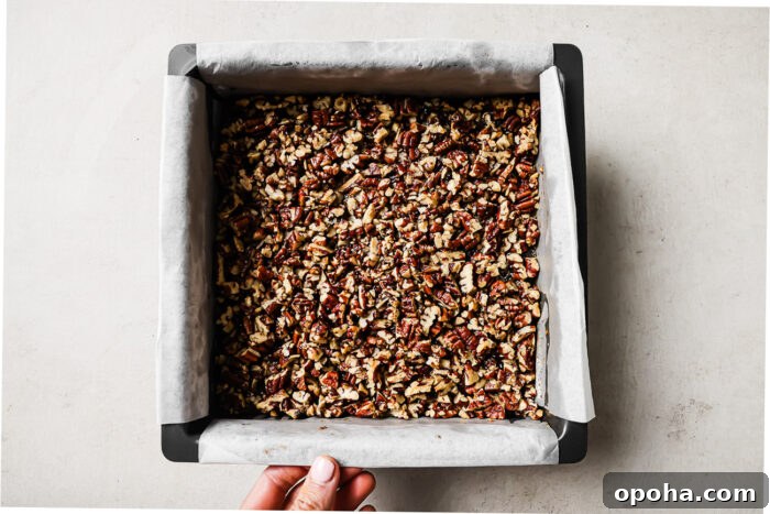 pecan pie brownies in a parchment-lined square baking pan, topped with chopped pecans.