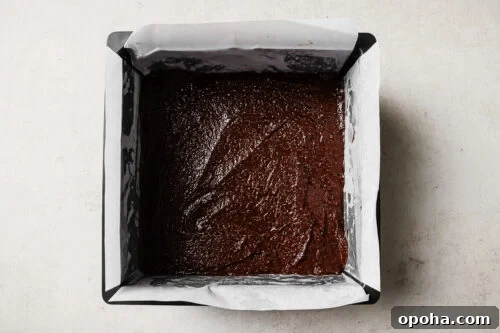 Chocolate brownie batter evenly spread in a parchment-lined square baking pan, ready to bake.