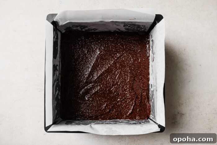 Chocolate brownie batter evenly spread in a parchment-lined square baking pan, ready to bake.