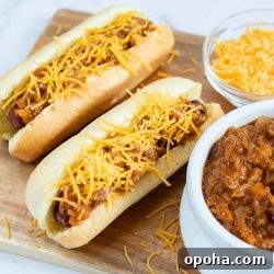 Loaded Chili Cheese Hot Dogs Recipe with Homemade Chili 2 Two hot dogs in buns topped with chili and shredded cheese, a bowl of shredded cheese, and a bowl of chili.