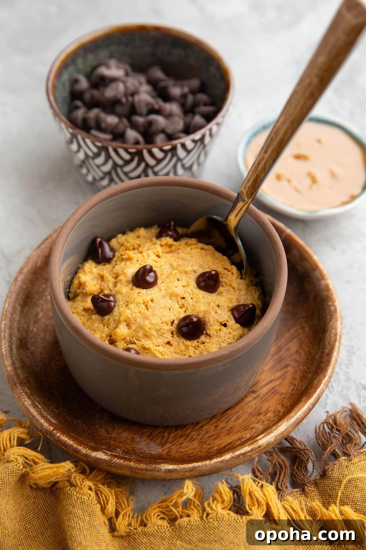 Peanut butter cake in a ramekin sitting on a wooden plate with a small bowl of peanut butter in the background and a bowl of chocolate chips.
