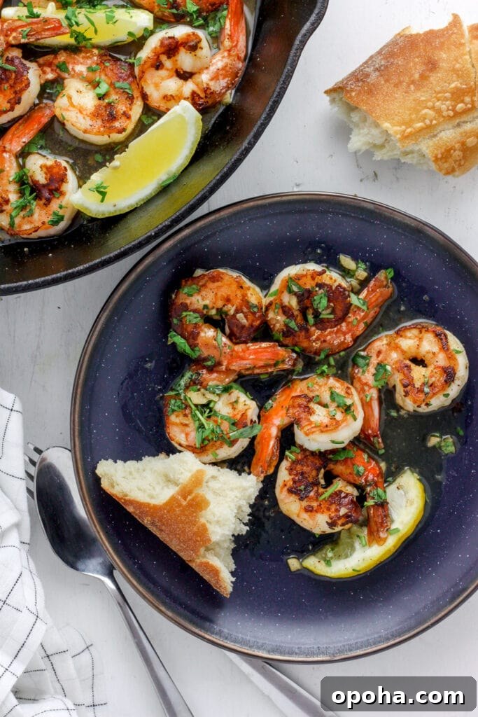 A blue plate with seared shrimp and crusty bread. off center is a skillet with more seared shrimp.