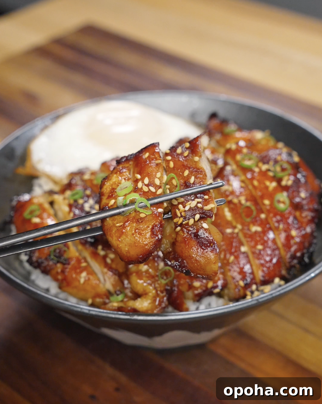Chopsticks lifting a piece of caramelized chicken bulgogi coated in a glossy gochujang-based sauce, topped with sesame seeds and sliced scallions over a bowl of rice.