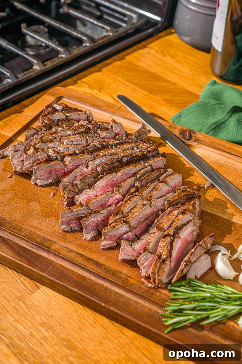 A wooden board with sliced London broil served with garlic and rosemary. There is a knife on the board.