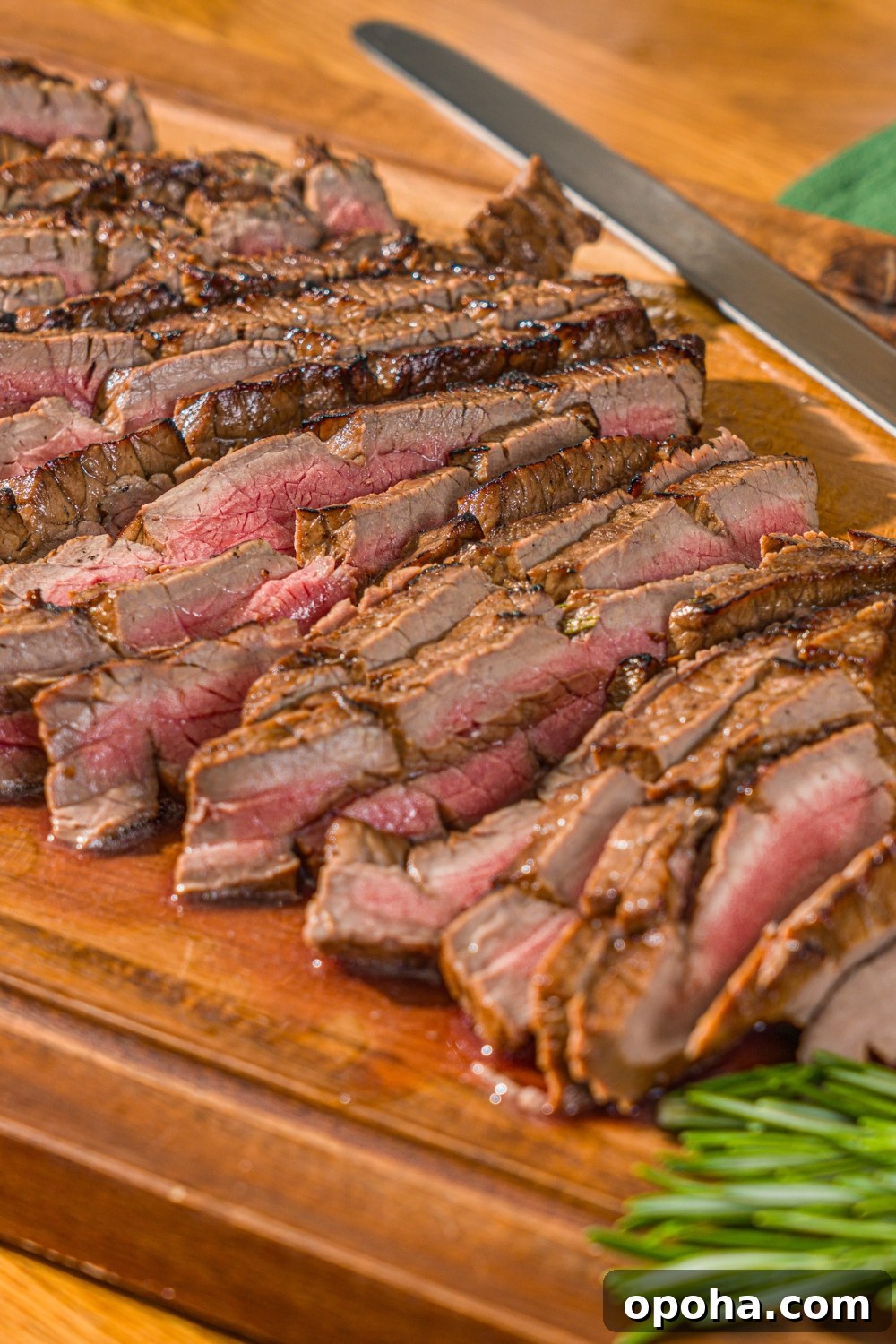 A wooden board with sliced London broil served with garlic and rosemary. There is a knife on the board.