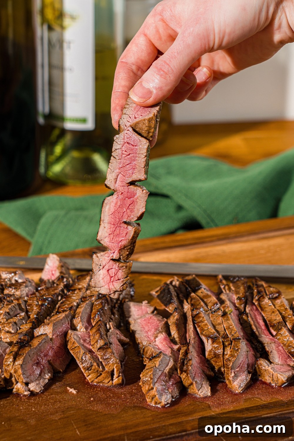 A wooden board with sliced London broil served with garlic and rosemary. A hand is holding a slice of beef.