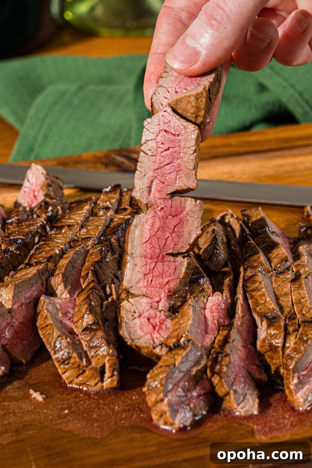 A wooden board with sliced London broil served with garlic and rosemary. A hand is holding a slice of beef.