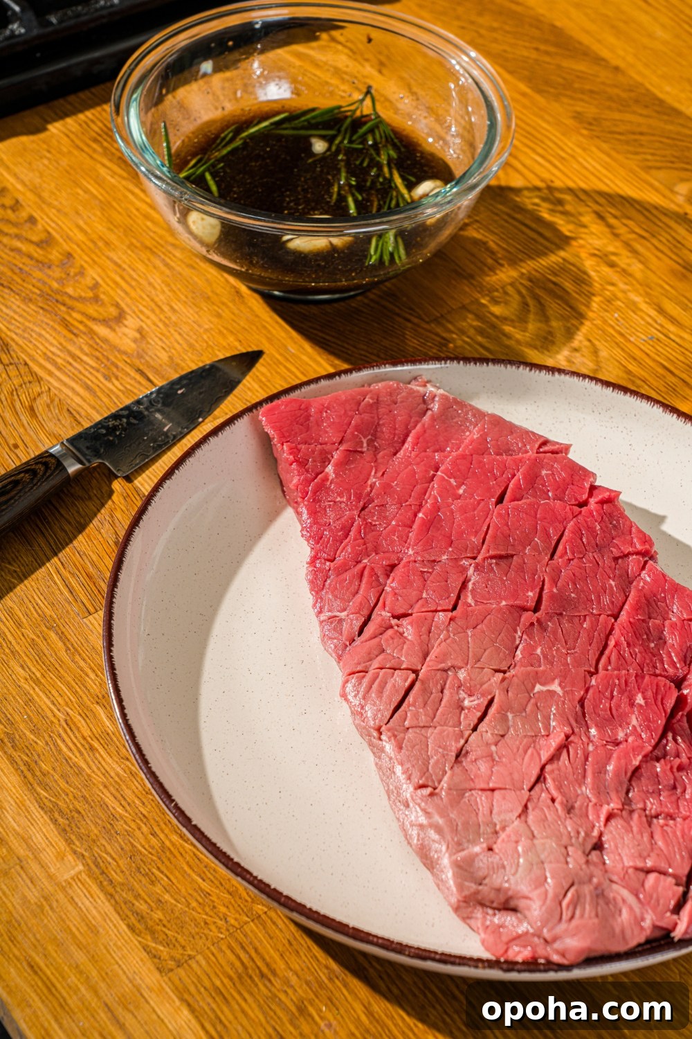 London broil beef scored in a diagonal pattern on a plate with a bowl of marinade.