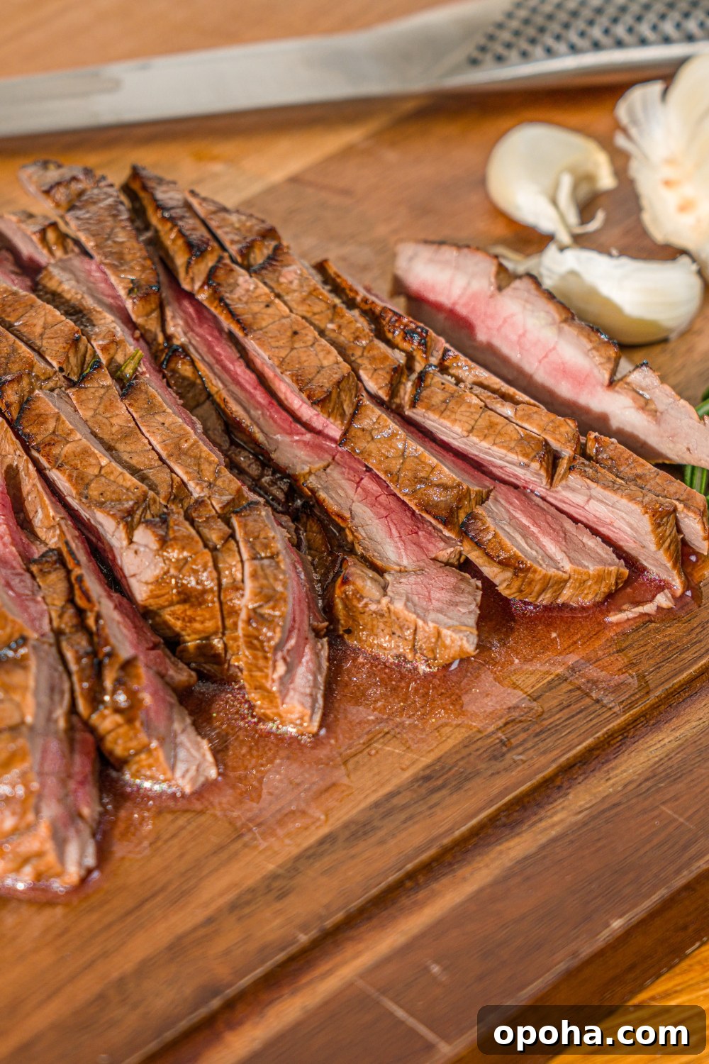 A wooden board with sliced London broil served with garlic and rosemary. There is a knife on the board.